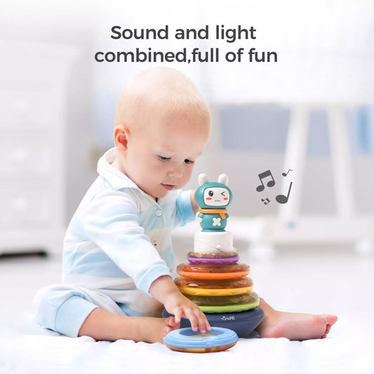 Baby sitting on a soft surface playing with a colorful stacking rings toy topped with a bunny character. The toy features multiple translucent rings in rainbow colors stacked on a wobbling base. Musical notes and text on the image highlight its sound and light functions with the caption: “Sound and light combined, full of fun.”