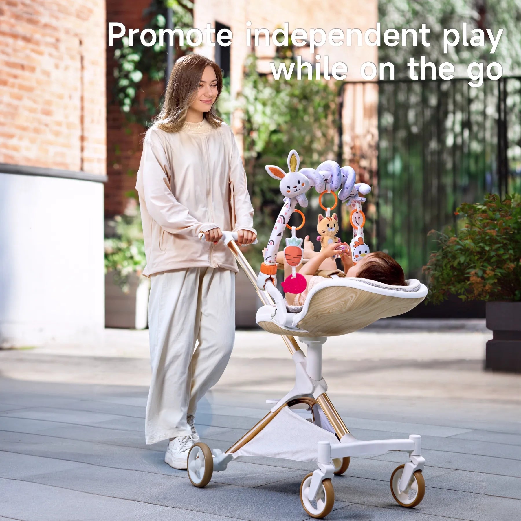 Mother pushing a stroller outdoors with a baby engaged in independent play, reaching for a colorful animal-themed hanging toy arch attached to the stroller.
