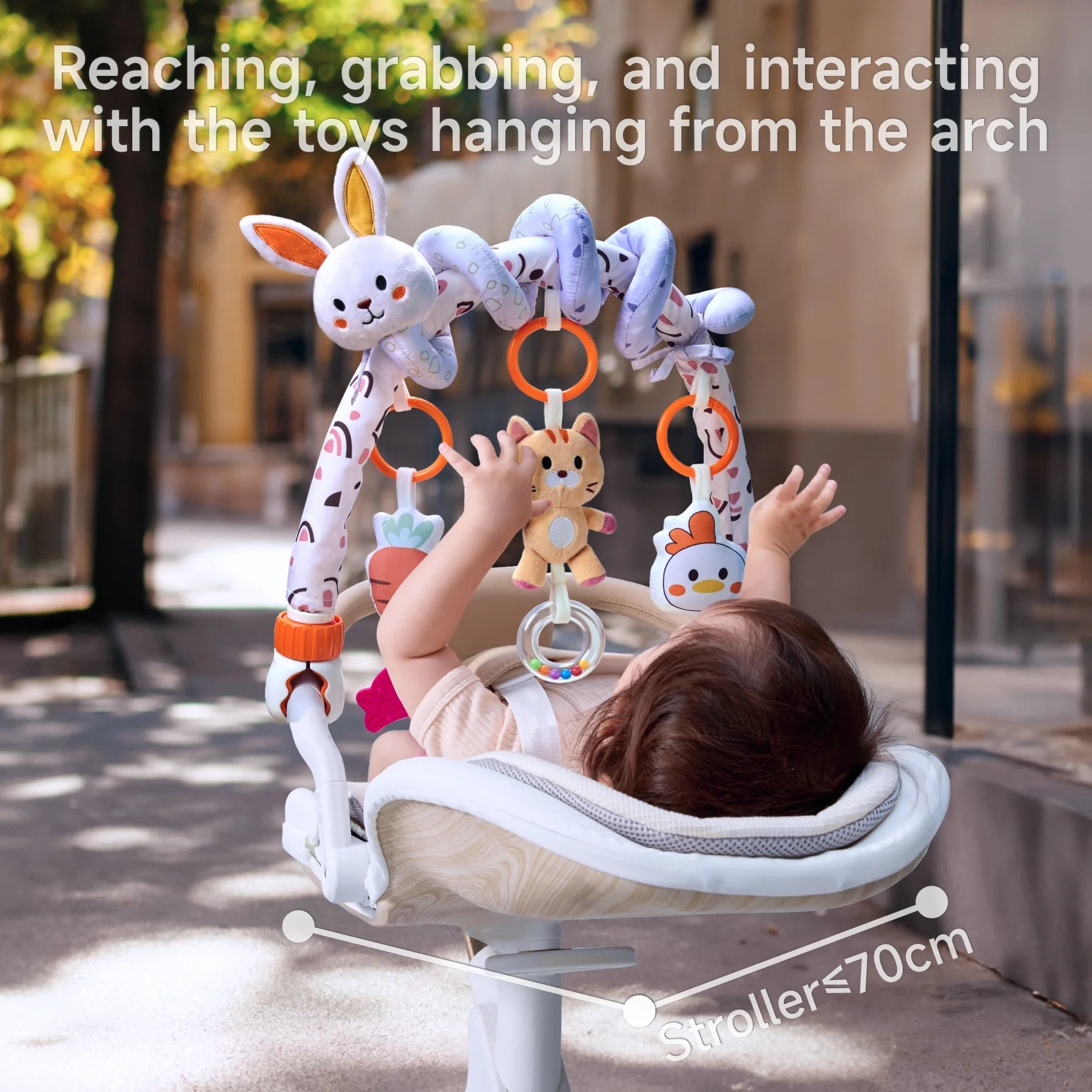 Baby lying in a stroller, actively reaching and grabbing plush animal toys hanging from a soft arch toy bar during an outdoor walk; suitable for strollers under 70cm wide.