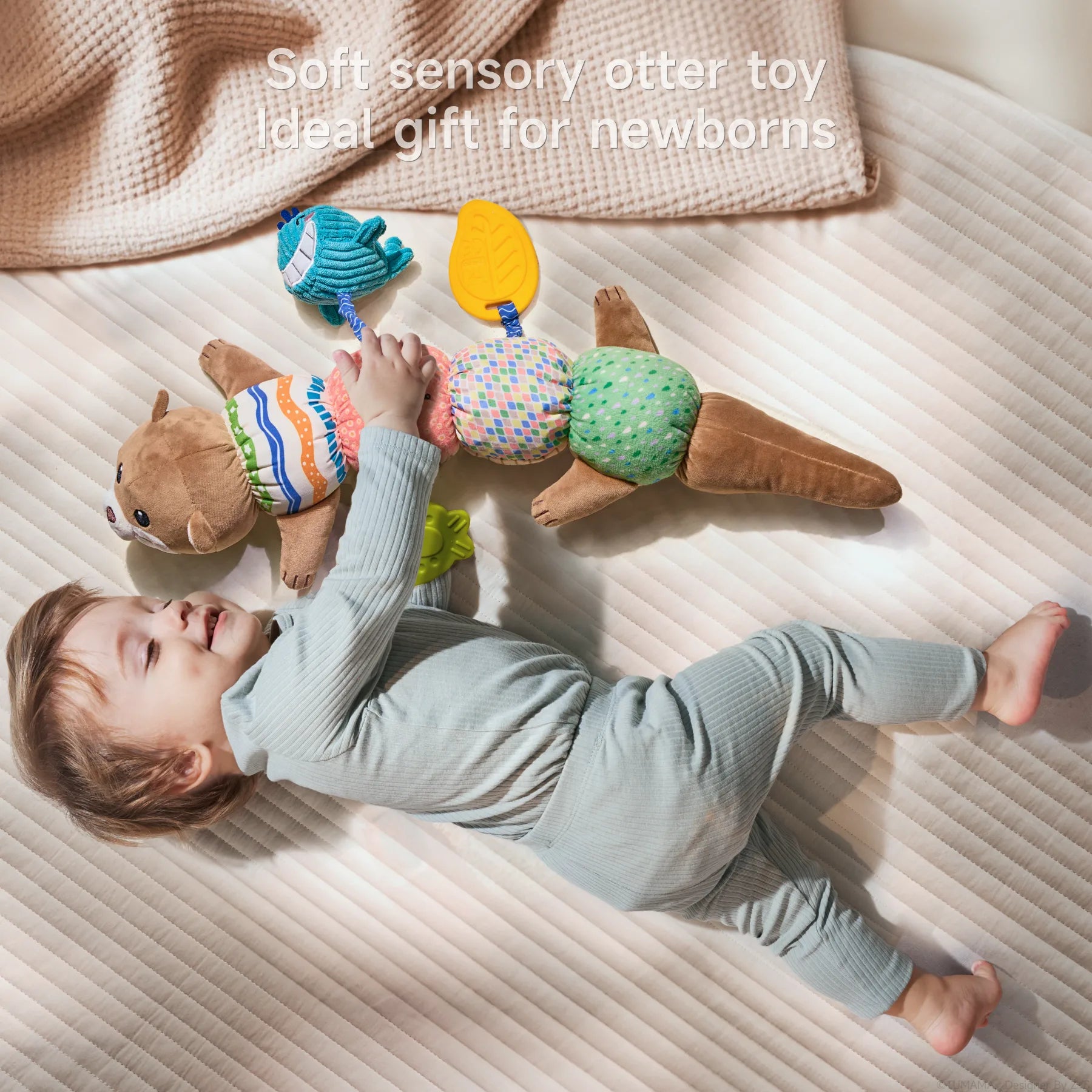 A joyful baby in light blue ribbed clothing lies on a cream-colored quilted surface, happily playing with a soft sensory otter toy. The plush toy features a caterpillar-like design with a brown otter head with cute facial features at one end and a tail at the other. Between them are five colorful textured segments in different patterns: striped, pink with circles, multicolored squares, and green with dots. Attached to the toy are a blue whale teether and a yellow leaf-shaped teether. Text overlay at the top