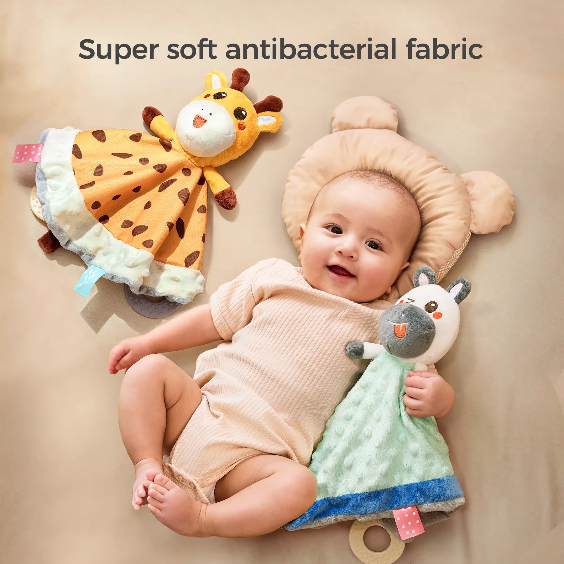 A smiling baby lying on a bed with a bear-shaped pillow, holding a plush zebra security blanket, while a plush giraffe security blanket lies nearby.