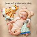A smiling baby lying on a bed with a bear-shaped pillow, holding a plush zebra security blanket, while a plush giraffe security blanket lies nearby.