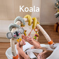 A baby lying in a bouncer, playing with a koala-themed arch toy featuring a plush koala, giraffe, bird, and star-shaped teether. The toy attaches securely, providing sensory and motor skill development fun.