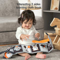 Baby sitting on the floor playing with a black and white double-sided cloth learning book, featuring cute animal illustrations and high-contrast patterns for early visual stimulation.