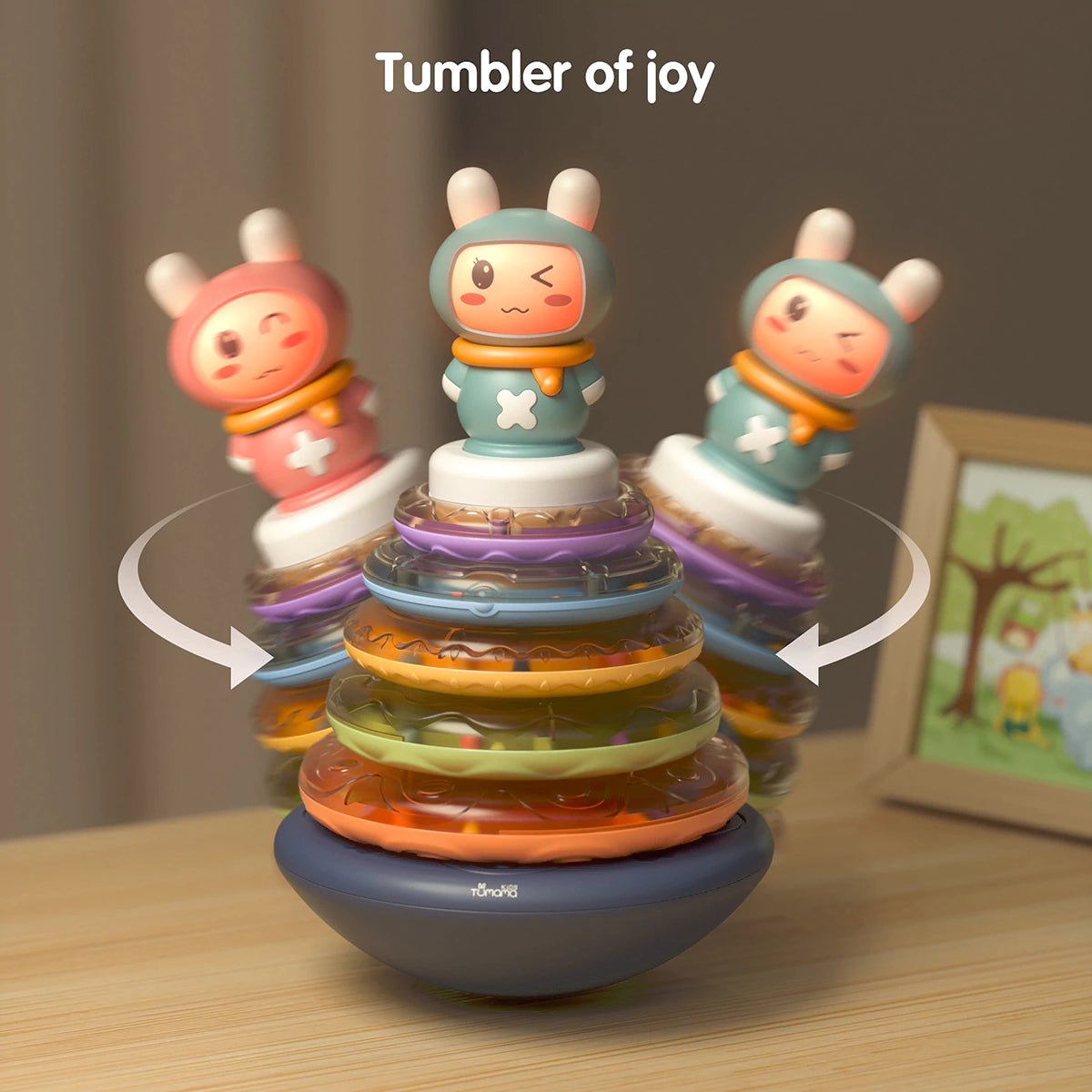 Musical stacking rings toy with a bunny topper wobbling side to side on its rounded base. The toy features translucent rainbow-colored rings stacked in layers, designed to rock back and forth like a tumbler. Motion arrows emphasize the wobbling action, and text at the top reads: “Tumbler of joy, get a lot of fun.”