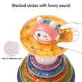 Close-up of a colorful stacking rings toy with a bunny topper. A hand is lifting one of the translucent orange rings filled with small beads, showing that each ring can also be used as a rattle. Text on the image reads: “Stacked with sound, but can be a rattle,” with an inset detail noting: “Play 10 different sound.”